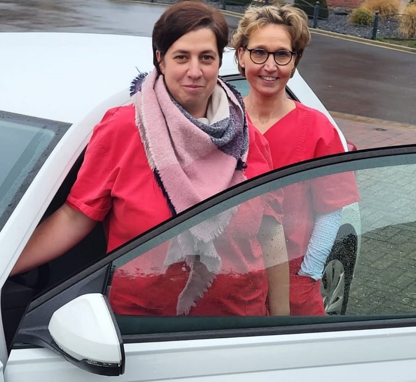 Zwei Frauen in roten Uniformen stehen neben einem Auto, eine von ihnen lächelt.