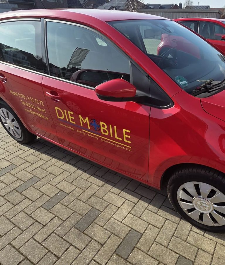 Zwei Frauen in roten Arztkitteln stehen neben einem weißen Auto mit Logo.